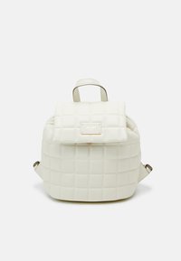 Sac à dos blanc matelassé avec un rabat, comprenant une plaque logo dorée et des bretelles réglables. Matériau lisse avec une forme structurée.