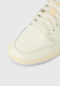 Cremfarbener Sneaker mit perforierter Lederzehe, sichtbaren Nähten, weißen Schnürsenkeln und einer hellen Gummisohle auf einem weißen Hintergrund.
