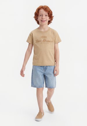 Garçon roux portant un t-shirt beige Levi Strauss, un short en denim bleu clair et des chaussures beige à enfiler, marchant vers l'avant sur un fond blanc.