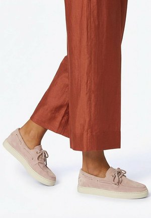 Person wearing rust-colored wide-leg pants and light pink suede lace-up sneakers, standing with one foot slightly raised.