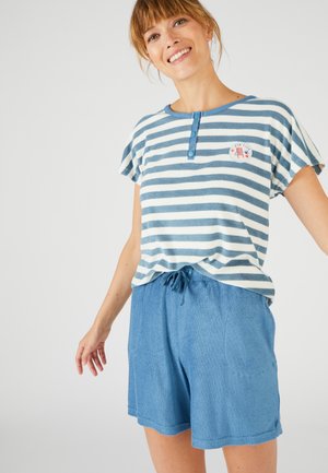 Glunderende vrouw die een blauw-wit gestreept shirt met korte mouwen en knopen draagt, en blauwe shorts met taillekoord, tegen een effen achtergrond.