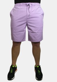 Pantaloni corti in cotone viola con elastico in vita, chiusura con cordino e tasche laterali. Indossati con scarpe da ginnastica nere con dettagli neon.