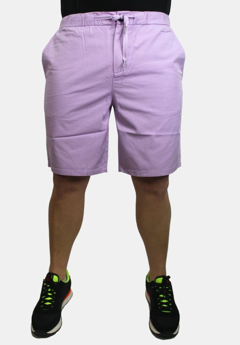 Pantaloni corti in cotone viola con elastico in vita, chiusura con cordino e tasche laterali. Indossati con scarpe da ginnastica nere con dettagli neon.