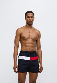 Μαύρο μαγιό βερμούδα με κόκκινη και λευκή οριζόντια ρίγα, διαθέτει ελαστική μέση με κορδόνι και ελαφρύ υλικό.