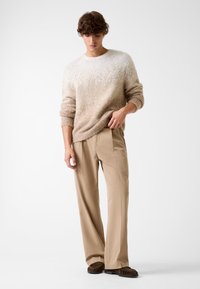 Maglione beige lavorato a maglia con una texture sfumata, abbinato a pantaloni beige a gamba larga e scarpe scamosciate marrone scuro. Palette di colori semplice e neutra.