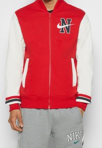 Chaqueta con cremallera en rojo y blanco, de textura suave, que presenta un logo negro en forma de "N" y un swoosh blanco, puños a rayas y dos bolsillos blancos.