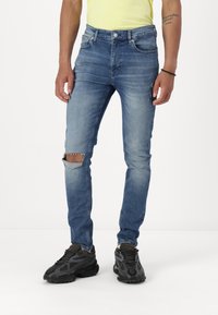 Jeans din denim albastru cu o croială slim, prezentând detalii uzate și o ripă la genunchi. Asortați cu adidași negri cu un design texturat.