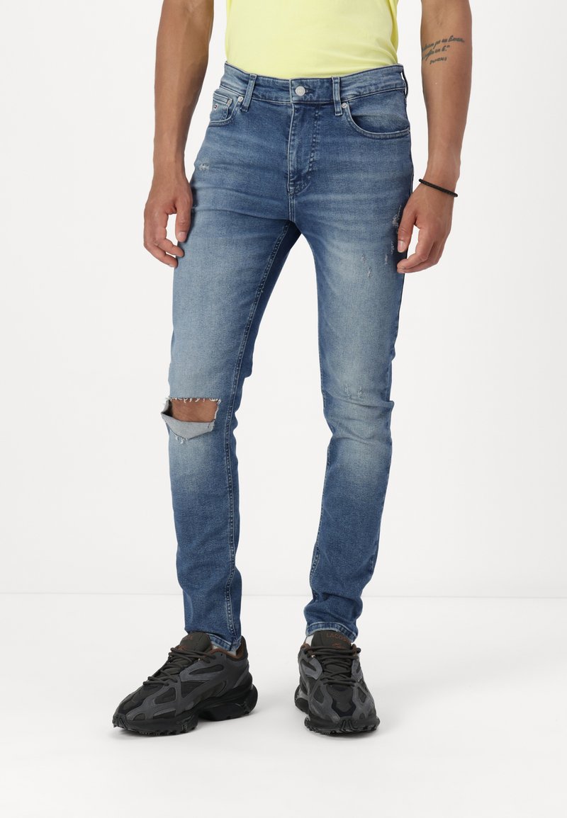 Jeans din denim albastru cu o croială slim, prezentând detalii uzate și o ripă la genunchi. Asortați cu adidași negri cu un design texturat.