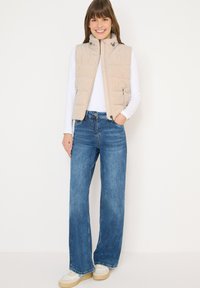 Chaleco acolchado beige con cuello alto, dos bolsillos laterales, usado sobre una camisa blanca de manga larga, combinado con jeans de pierna ancha azul claro y zapatillas deportivas.