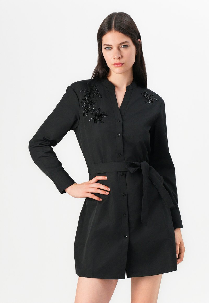 Schwarzes Hemdkleid mit langenÄrmeln, Knopfverschluss und Taillengürtel. Verfügt über detaillierte schwarze Perlenstickerei auf den Schultern. Glattes, maßgeschneidertes Gewebe.