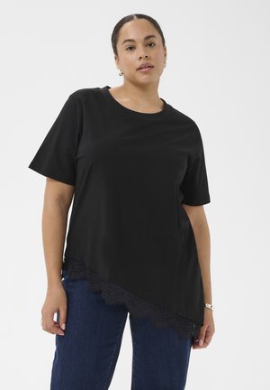 Kvinde iført en sort kortærmet top med asymmetrisk blonderkant og mørkeblå jeans, stående mod en ensfarvet hvid baggrund.