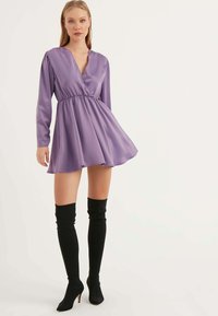 Lila Satin-Minikleid mit langen Ärmeln, V-Ausschnitt und geraffter Taille; kombiniert mit schwarzen Overkneestiefeln. Glatte Textur, ausgestellter Rock.