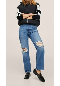 Schwarze Daunenjacke mit weißen gestreiften Ärmeln, kombiniert mit ausgewaschenen blauen Jeans mit großen Rissen. Schwarze Slipper runden das Outfit ab.