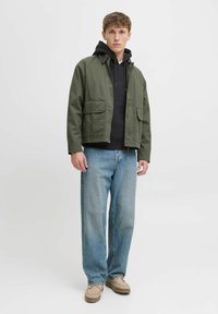 Giacca verde oliva, felpa nera sotto, jeans larghi azzurri chiari, calzature beige. Due tasche frontali sulla giacca, chiusura con bottoni, stile casual.