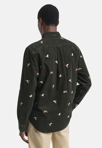 Man in een donkergroene langemouwshirt met geborduurde vliegende eenden, beige broek, gezien vanaf de achterkant tegen een effen achtergrond.