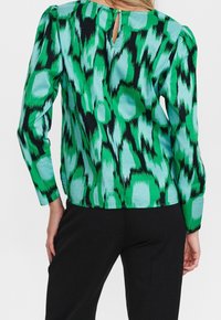 Blouse à motifs verts et noirs avec des manches longues, présentant un détail de décolleté en trou de serrure au dos. Tissu doux avec une texture lisse.