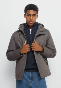 Szara kurtka z kapturem, zapinana na zamek i napy, z bocznymi kieszeniami, a pod nią ma teksturowany granatowy sweter, na białym tle.