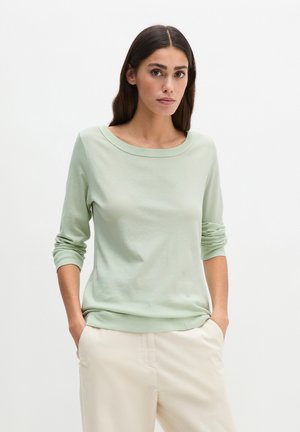 Femme aux longs cheveux noirs portant un haut à manches longues vert clair et un pantalon crème avec les mains dans les poches, sur un fond uni.