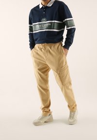 Granatowa koszulka polo z długim rękawem, białym kołnierzykiem, zielonym paskiem i nadrukowanym logo, zestawiona z beżowymi spodniami typu cargo z bocznymi kieszeniami i elastycznym pasem.
