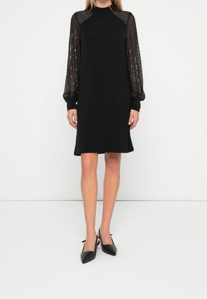 Femme portant une robe noire midi avec des manches longues transparentes ornées de perles et des escarpins noirs pointus à bride arrière, debout devant un fond blanc.