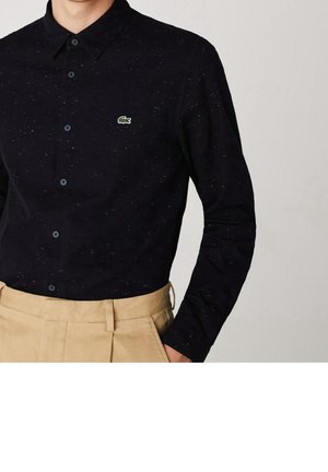 Uomo che indossa una camicia nera a maniche lunghe con bottoni e macchie, con un piccolo logo di un coccodrillo verde, e pantaloni beige a vita alta.