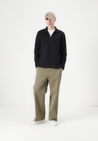 Junger Mann steht mit Händen in den Taschen, trägt eine beige Kappe, einen schwarzen Halbzip-Pullover, khakifarbene Hose und weiße Sneaker vor einem einfarbigen Hintergrund.