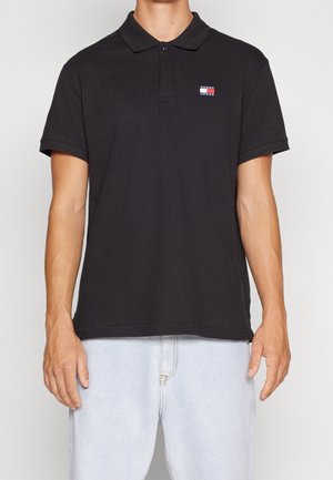 Homme portant un polo noir avec un petit logo Tommy Jeans sur la poitrine et un jean bleu clair, se tenant devant un fond uni.