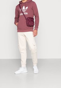 Bordeaux huva med vitt logotyp, krämfärgade joggers med cargo-fickor och vita sneakers. Inkluderar en liten burgundy axelväska.