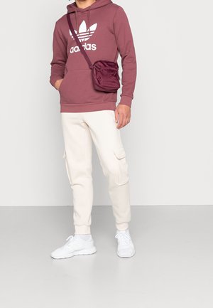 Bordeaux hoodie met wit logo, crèmekleurige joggers met cargozakken en witte sneakers. Inclusief kleine bordeaux schoudertas.