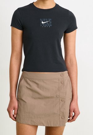 Kobieta ubrana w czarny krótki T-shirt Nike z logo i beżową mini spódnicę na guziki, stojąca z rękami swobodnie opuszczonymi wzdłuż ciała.