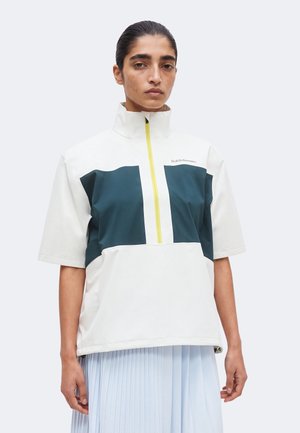 Άτομο που φοράει λευκό και σκούρο τυρκουάζ κοντομάνικο μπουφάν με κίτρινο φερμουάρ και ανοιχτό μπλε πλισέ φούστα σε απλό φόντο.