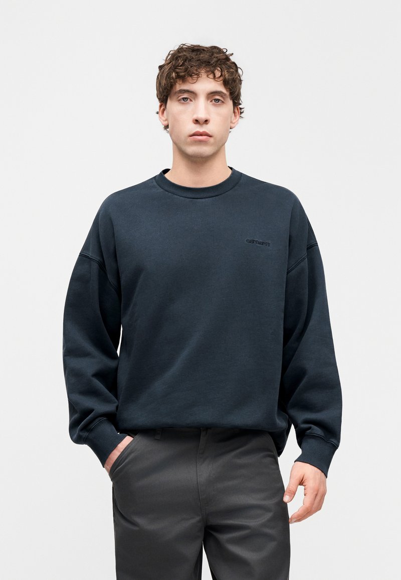 Felpa blu scuro con collo rotondo, polsini a costine e un piccolo logo nero sul petto; abbinata a pantaloni grigi.