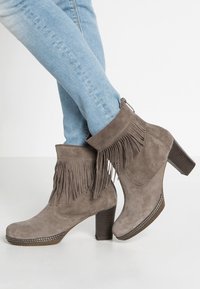 Bottines en suède gris avec des accents à franges, un talon texturé et un bout rond, associées à un jean bleu clair sur un fond blanc.