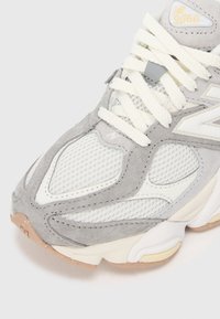 Baskets New Balance 9060 gris et blanches avec des panneaux en mesh et en suède, des lacets crème et une semelle beige sur un fond clair.