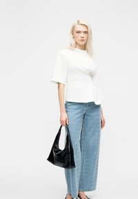 Νέα γυναίκα με άσπρο κοντομάνικο τοπ και μπλε τζιν με σχέδια, κρατώντας μια μαύρη τσάντα, στέκεται μπροστά σε απλό λευκό φόντο.