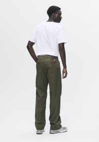 Olive green cargo pants with two back pockets, featuring a small red logo. Worn with a white t-shirt and gray sneakers.