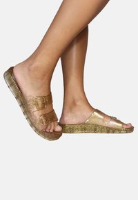 Sandales dorées pailletées avec deux boucles réglables, fabriquées en plastique brillant, dotées d'une semelle plate et d'un design à bout ouvert.