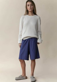 Witte geribde trui, blauwe polka dot short, en grijze klompen. Gelaagd stof, losse pasvorm en casual stijl met verlaagde schoudernaden.