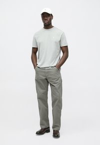 T-shirt de algodão cinzento claro com pequeno logótipo, combinada com calças cargo verde oliva e botas de couro pretas. Boné branco completa o conjunto.