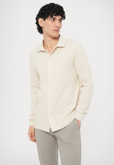 Πουκάμισο με κουμπιά και μακριά μανίκια σε μπεζ ύφασμα με υφασμένο διαμαντένιο μοτίβο, κλασικό γιακά και μανίκια με μανσέτες· συνδυασμένο με γκρι παντελόνι.