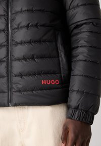 Zwarte gewatteerde jas met horizontale stiksels, ritssluiting en rode "HUGO" branding aan de linkerkant. Elastische manchetten en een gladde textuur.