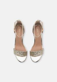 Högklackade sandaler med remmar i metalliskt silver och ett glittrande guldband över ovandelen. Har en ankelrem med spänne.
