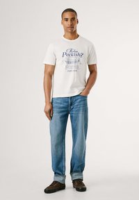 Camiseta de algodón blanca con texto gráfico azul "¿Tienes hambre?" que presenta una ilustración de un sándwich. Combinada con jeans de mezclilla azul claro y zapatos marrones.