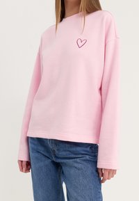 Sudadera rosa de corte relajado, con mangas largas y un pequeño detalle bordado de un corazón en el pecho. Material de algodón texturizado.
