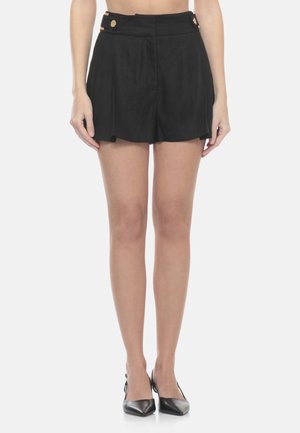 Donna che indossa shorts neri a vita alta con bottoni laterali e décolleté nere con punta affilata, in piedi davanti a uno sfondo bianco neutro.