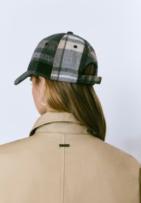 Gorra de cuadros gris y negra en mezcla de lana con visera curva y correa trasera ajustable, llevada con una chaqueta beige con cuello.