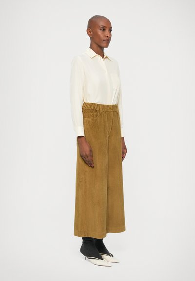 Camisa de manga larga color crema con un bolsillo, combinada con pantalones de pana amarillos de tiro alto y acanalados, y tacones blancos de punta.