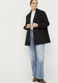 Zwarte wollen jas met een reverskraag en twee knopen, gedragen over een witte coltrui, gecombineerd met lichtblauwe, wijde jeans en bruine laarzen.