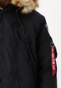 Black parka jacket with a fur-lined hood, multiple zippered pockets, and a red tag reading "REMOVE BEFORE FLIGHT." Textured fabric, utilitarian design.