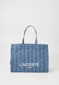 Mėlyna audinio rankin�ė su geometriniu raštu, turinti dvi rankenas ir didelį baltą "LACOSTE 1933" logotipą priekyje.